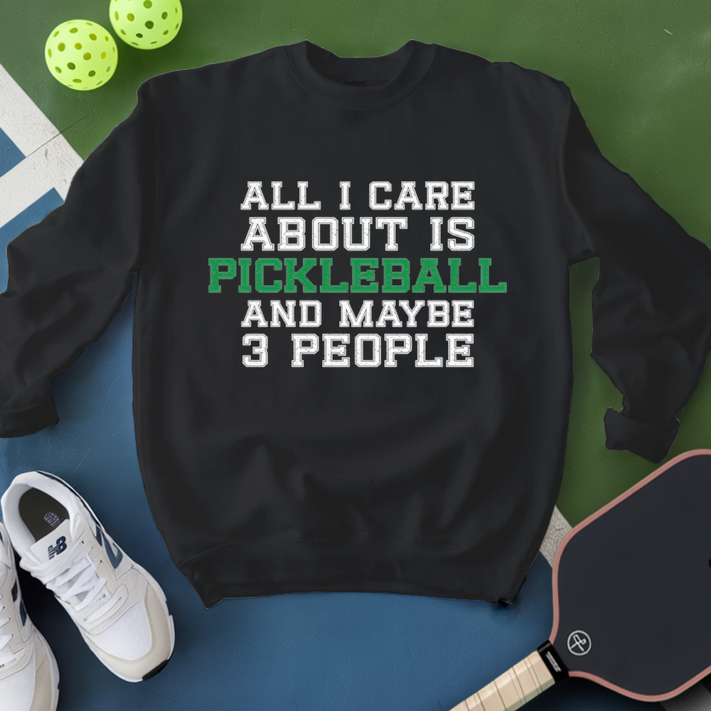 Black sweatshirt with pickleball text, surrounded by pickleballs and a paddle on a green and blue background.