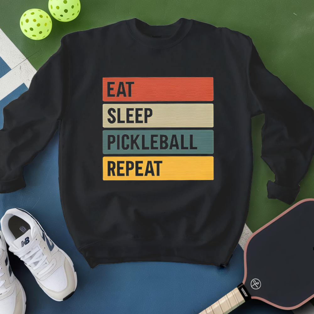 Black sweatshirt with 'Eat Sleep Pickleball Repeat' text, surrounded by pickleballs and a paddle on a green and blue background.