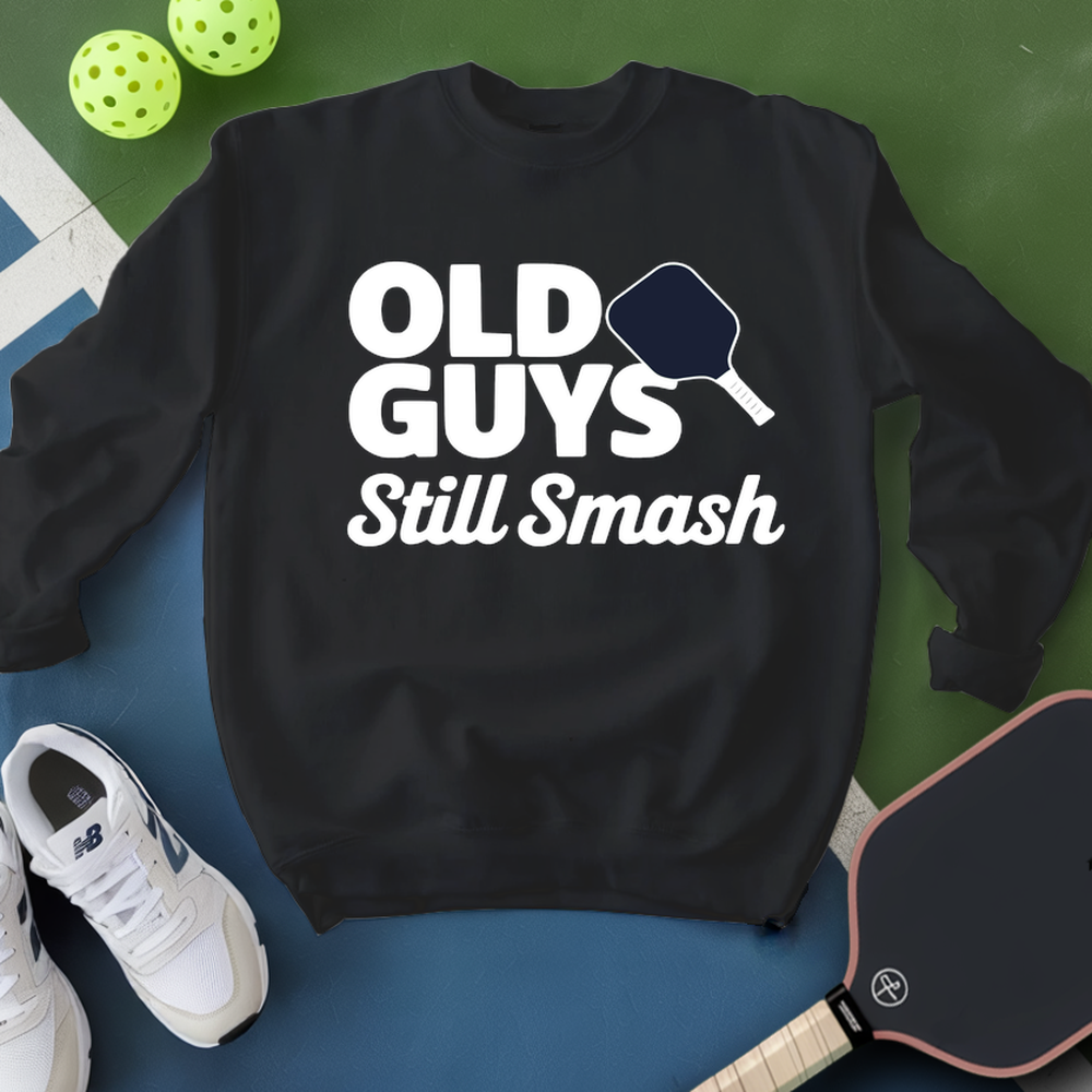 Black sweatshirt with 'Old Guys Still Smash' text, white sneakers, and pickleball paddle on a green and blue background