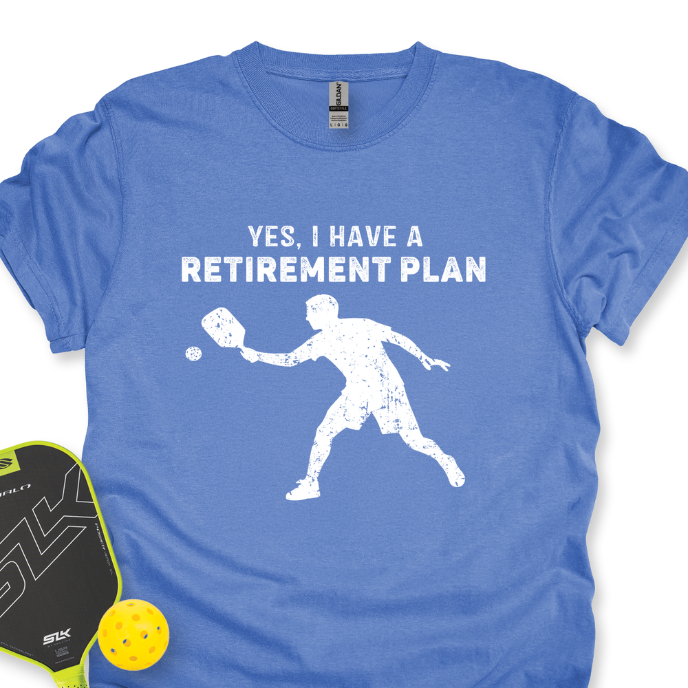 Blue t-shirt with 'Yes, I have a retirement plan' text and pickleball player graphic, accompanied by pickleball paddle and ball on a white background.