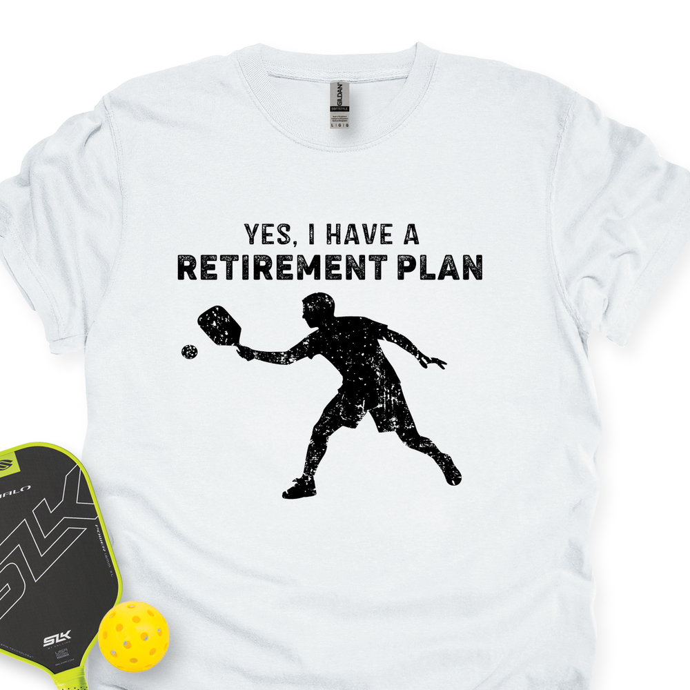White t-shirt with black graphic and text 'Yes, I have a retirement plan' next to pickleball equipment on a white background