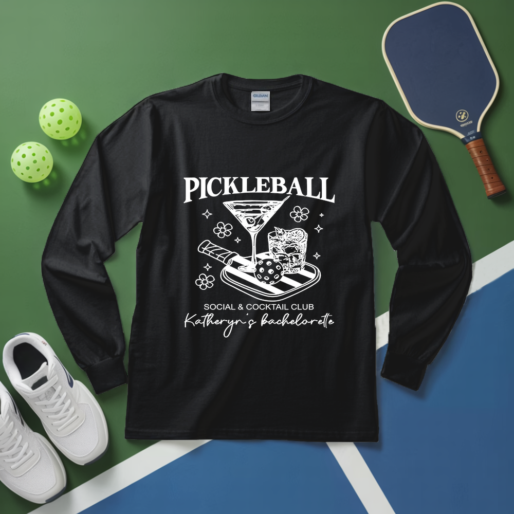Black long-sleeve shirt with pickleball-themed design on a green and blue surface with pickleballs and a paddle.