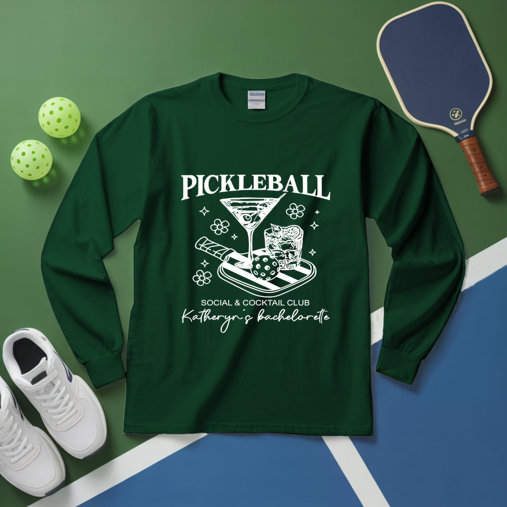 Green long-sleeve shirt with pickleball-themed design on a green and blue surface with pickleballs and a paddle.