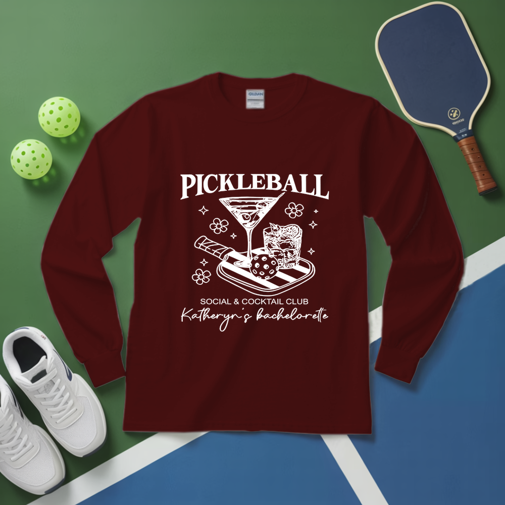 Maroon long-sleeve shirt with pickleball design on a green and blue surface with pickleballs and a paddle.