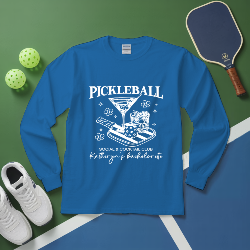 Blue long-sleeve shirt with pickleball design on a green and blue surface with pickleballs and paddle.