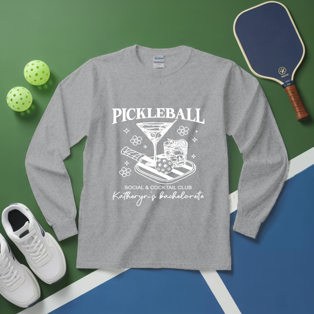Gray long-sleeve shirt with pickleball-themed design on a green and blue surface with pickleballs and a paddle.