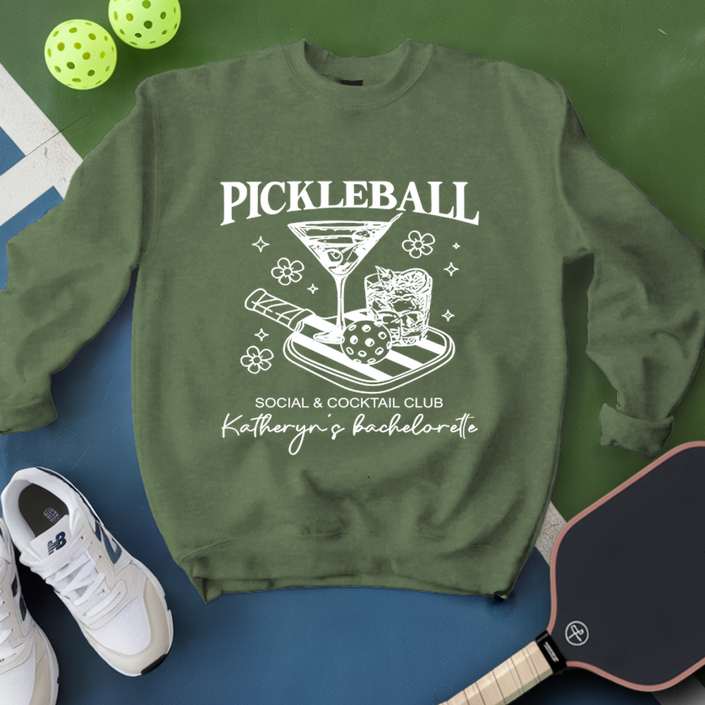 Green sweatshirt with pickleball design and text, placed on a blue surface with pickleballs and a paddle.