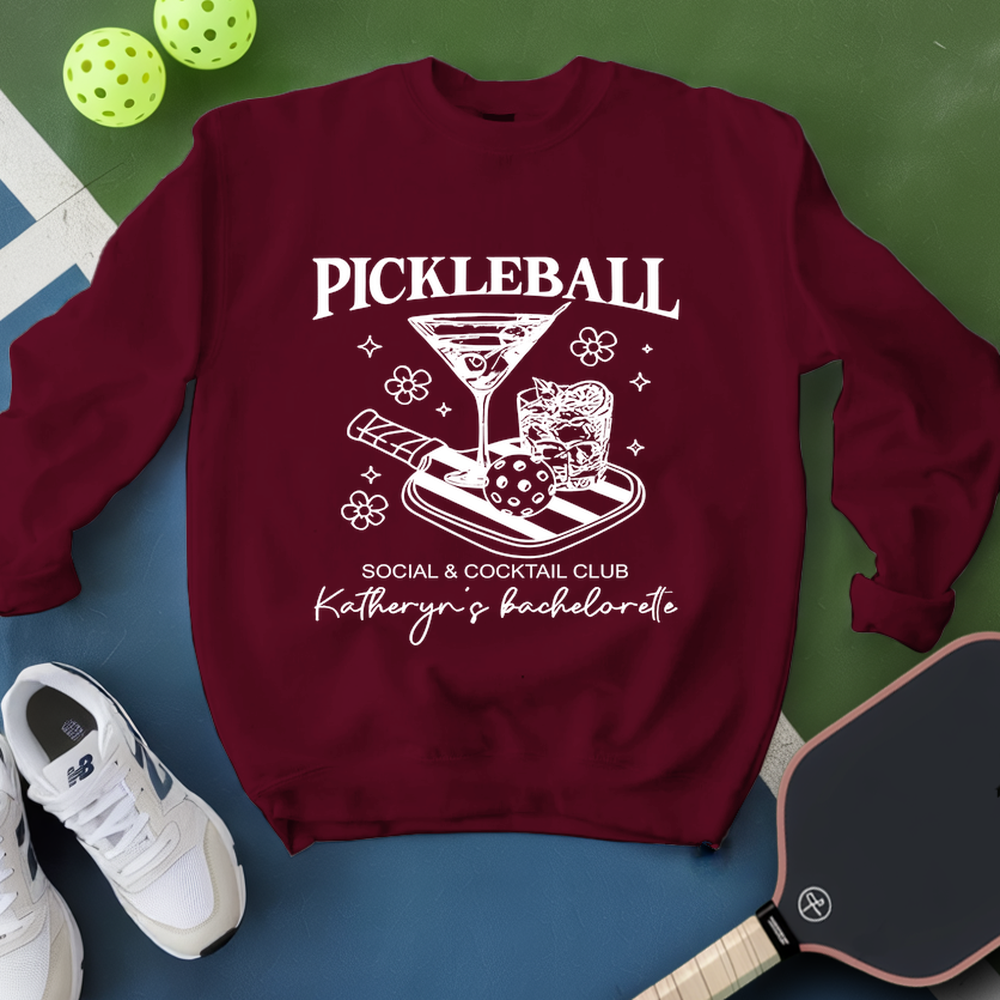 Maroon sweatshirt with pickleball design and text, placed on a blue and green surface with sports equipment.