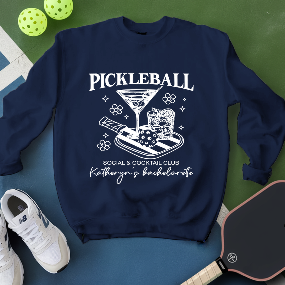 Navy blue sweatshirt with pickleball-themed design on a green and blue surface with pickleballs and a paddle.