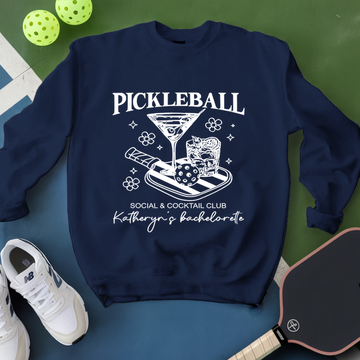 Navy blue sweatshirt with pickleball-themed design on a green and blue surface with pickleballs and a paddle.