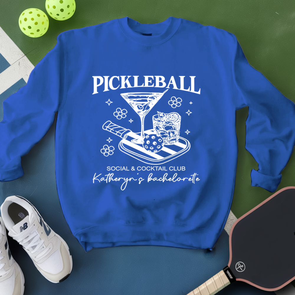 Blue sweatshirt with pickleball-themed design on a green surface with pickleballs and a paddle.