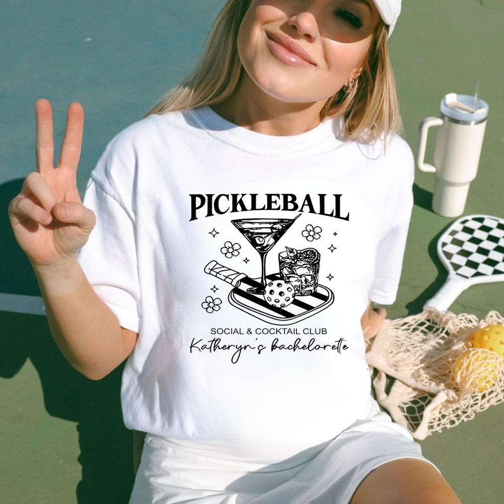 Person wearing a white t-shirt with 'Pickleball Social & Cocktail Club' design, sitting outdoors.