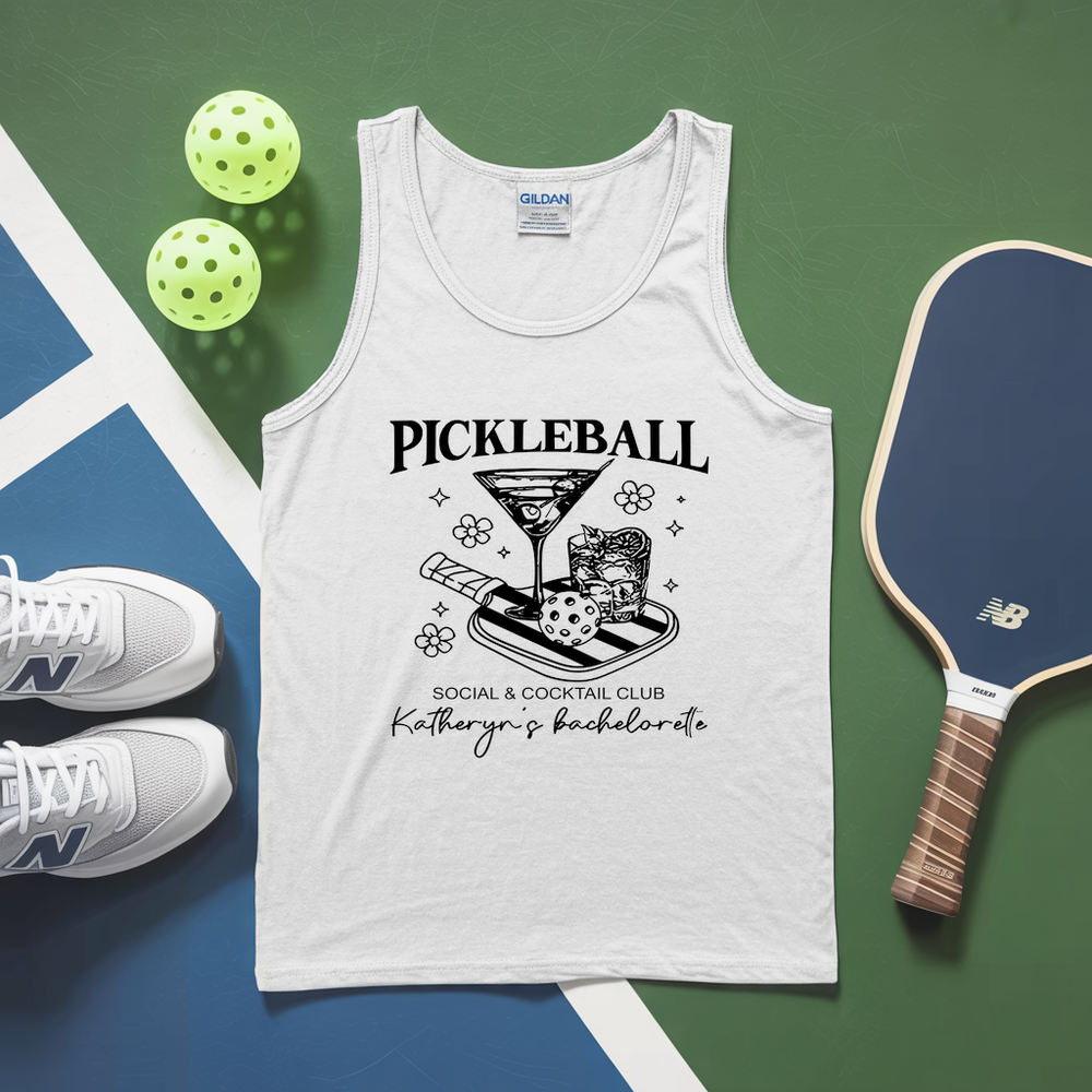 White tank top with pickleball-themed design on a green and blue surface with pickleballs and a paddle.