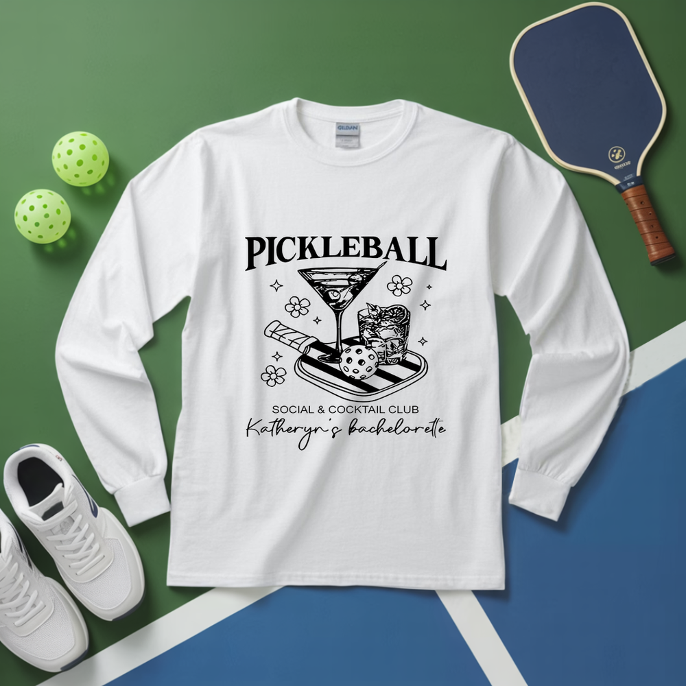 White long-sleeve shirt with pickleball-themed design on a green and blue surface with pickleballs and a paddle.