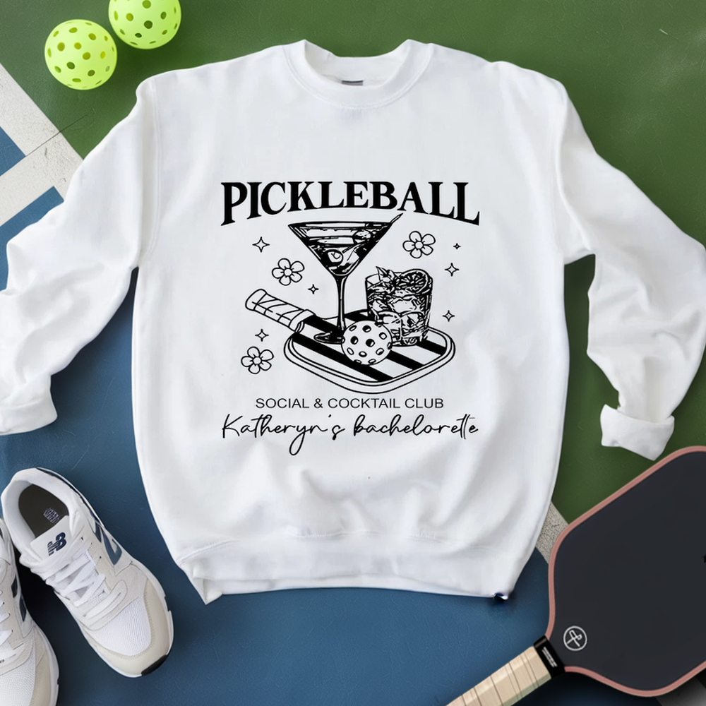 White sweatshirt with pickleball-themed design on a green and blue surface with pickleballs and a paddle.