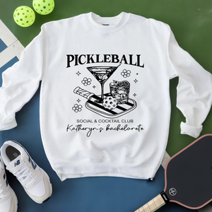 White sweatshirt with pickleball-themed design on a green and blue surface with pickleballs and a paddle.