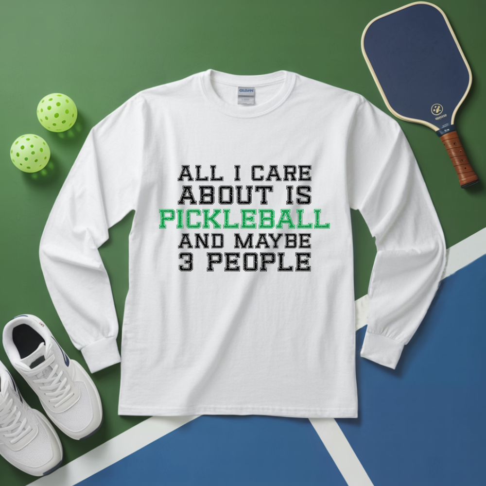 White long-sleeve shirt with pickleball-themed text on a green and blue surface with pickleball equipment.