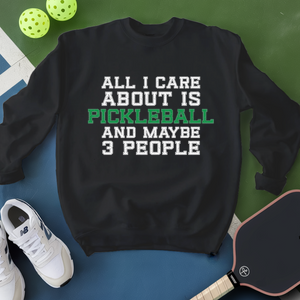 Black sweatshirt with pickleball text, surrounded by pickleballs and a paddle on a green and blue background.
