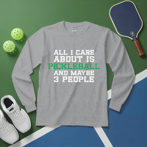 Gray long-sleeve shirt with pickleball-themed text on a green and blue surface with pickleball equipment.