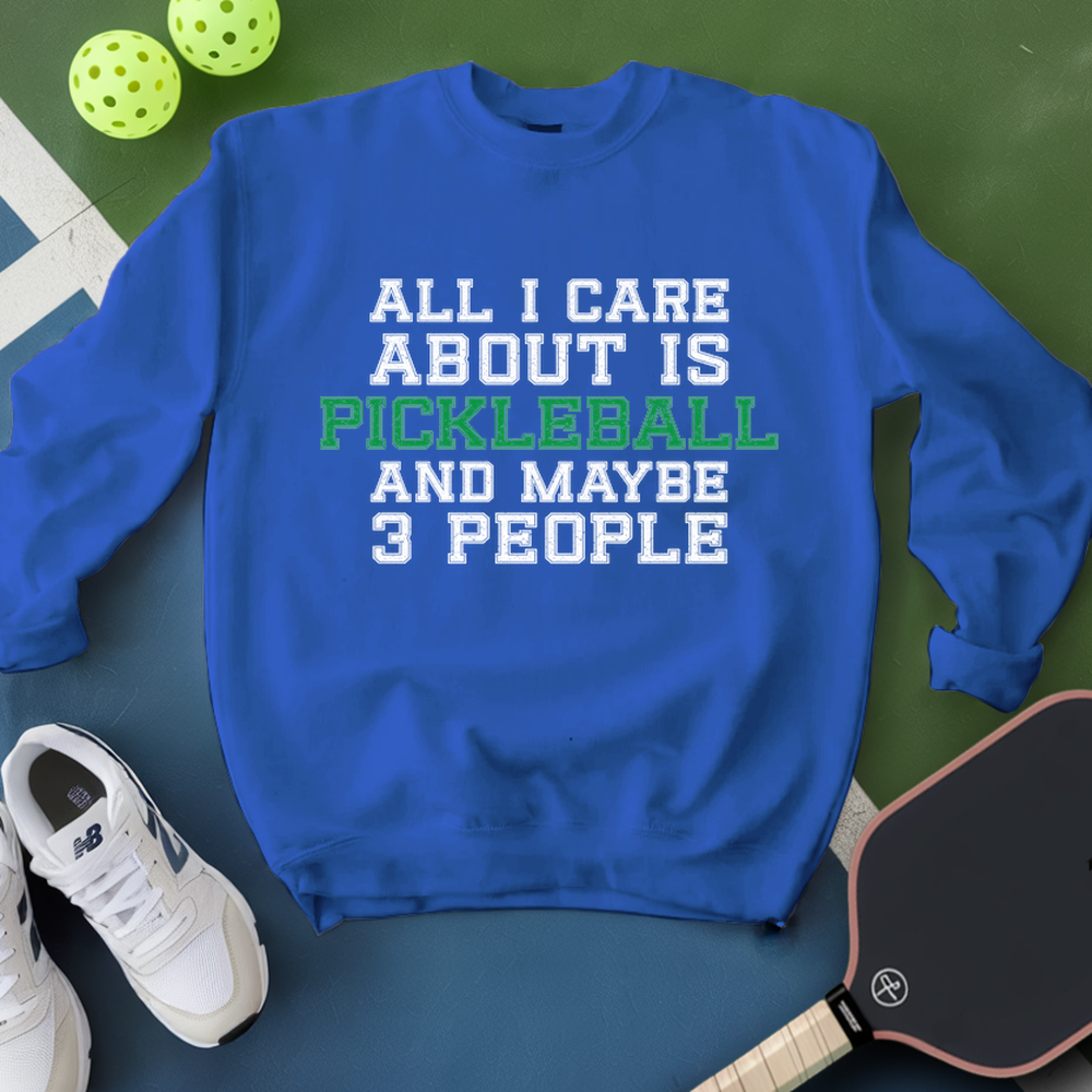 Blue sweatshirt with text 'All I care about is pickleball and maybe 3 people' on a green surface with pickleballs and a paddle.
