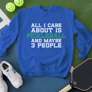 Blue sweatshirt with text 'All I care about is pickleball and maybe 3 people' on a green surface with pickleballs and a paddle.