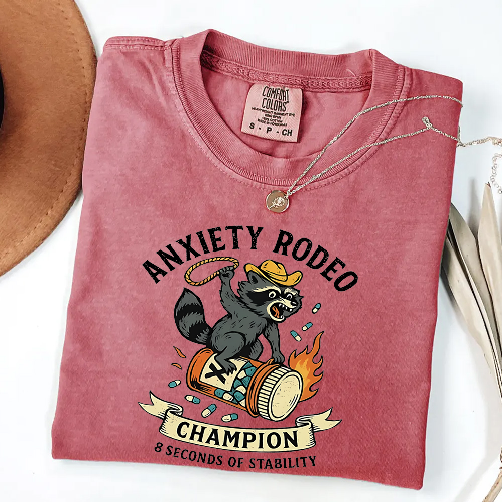 Folded pink t-shirt with 'Anxiety Rodeo Champion' graphic design on a white background