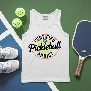 White tank top with 'Certified Pickleball Addict' design, pickleballs, and paddle on a green and blue background.