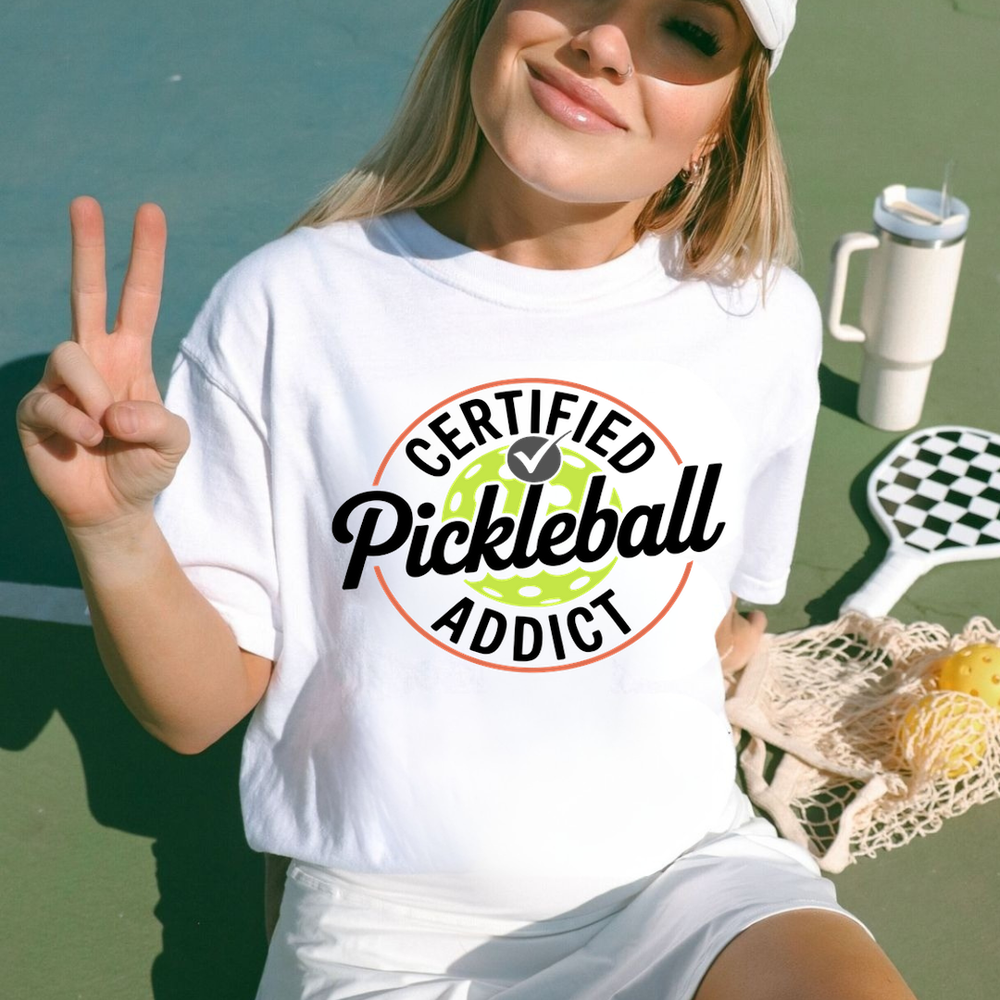 Person wearing a 'Certified Pickleball Addict' shirt on a pickleball court.