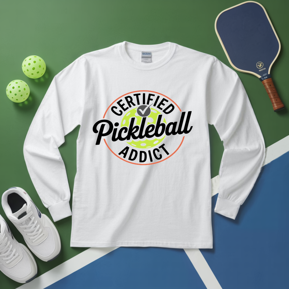 White long-sleeve shirt with 'Certified Pickleball Addict' design on a green and blue surface with pickleball equipment.