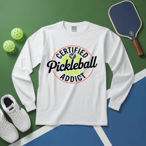White long-sleeve shirt with 'Certified Pickleball Addict' design on a green and blue surface with pickleball equipment.