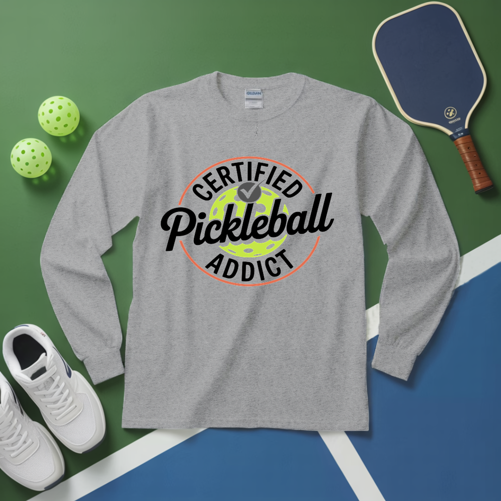 Gray long-sleeve shirt with 'Certified Pickleball Addict' design on a green and blue surface with pickleball equipment.
