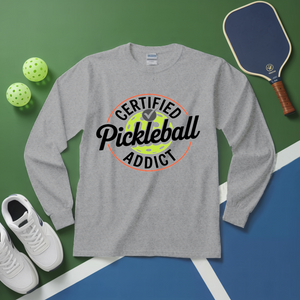 Gray long-sleeve shirt with 'Certified Pickleball Addict' design on a green and blue surface with pickleball equipment.