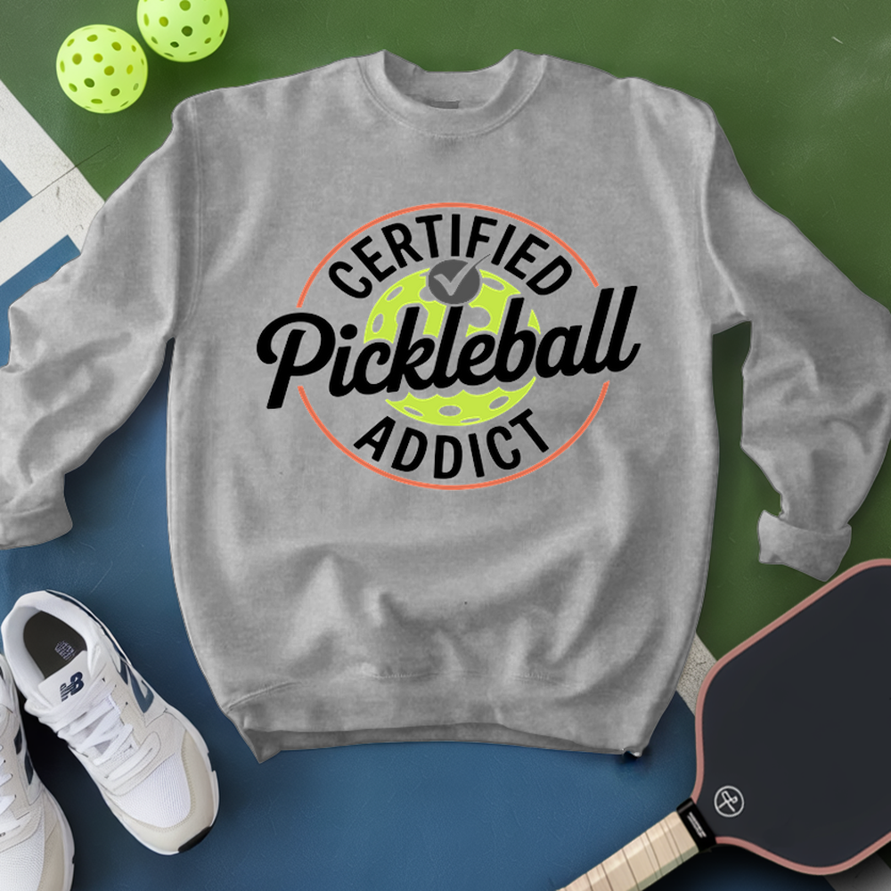 Gray sweatshirt with 'Certified Pickleball Addict' text, pickleball ball, paddle, and shoes on a green and blue background