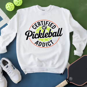 White sweatshirt with 'Certified Pickleball Addict' text, pickleball paddle, and balls on a green and blue background