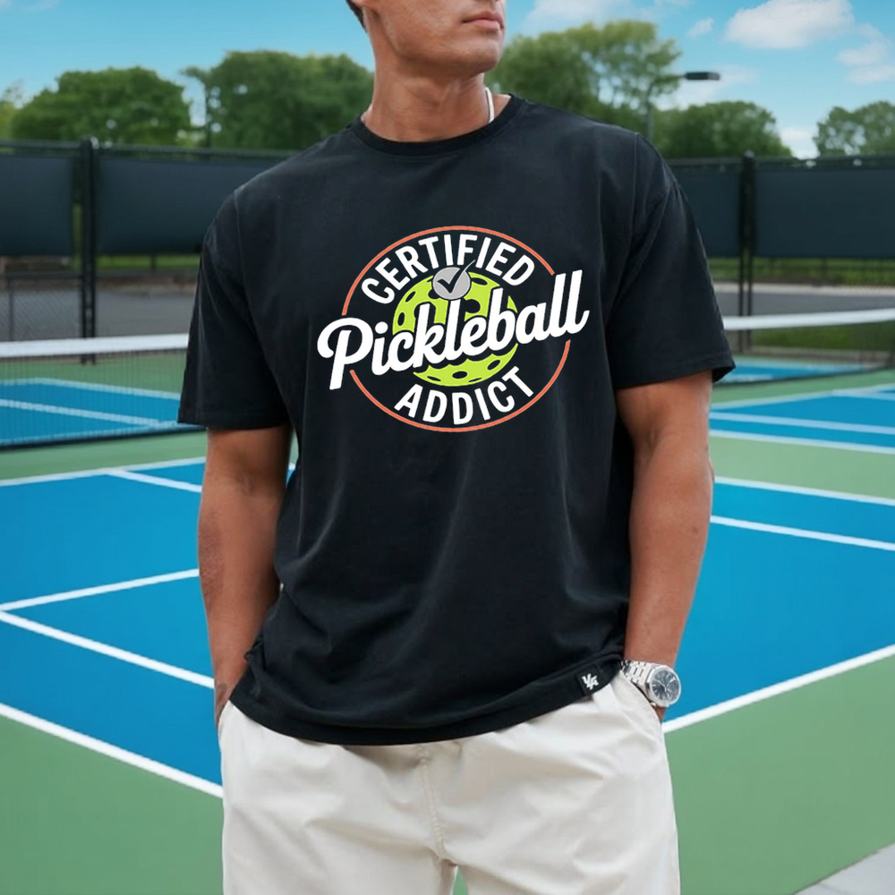 Person wearing a black t-shirt with 'Certified Pickleball Addict' text on a pickleball court.