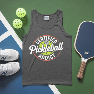 Gray tank top with 'Certified Pickleball Addict' design, pickleball balls, paddle, and shoes on a green and blue surface.