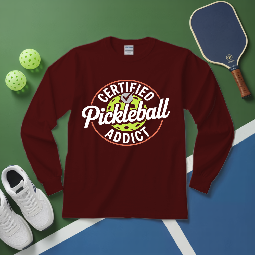 Maroon long-sleeve shirt with 'Certified Pickleball Addict' design on a green and blue surface with pickleball equipment.