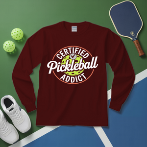 Maroon long-sleeve shirt with 'Certified Pickleball Addict' design on a green and blue surface with pickleball equipment.