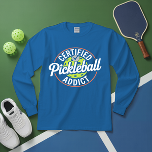 Blue long-sleeve shirt with 'Certified Pickleball Addict' design on a green and blue surface with pickleball equipment.
