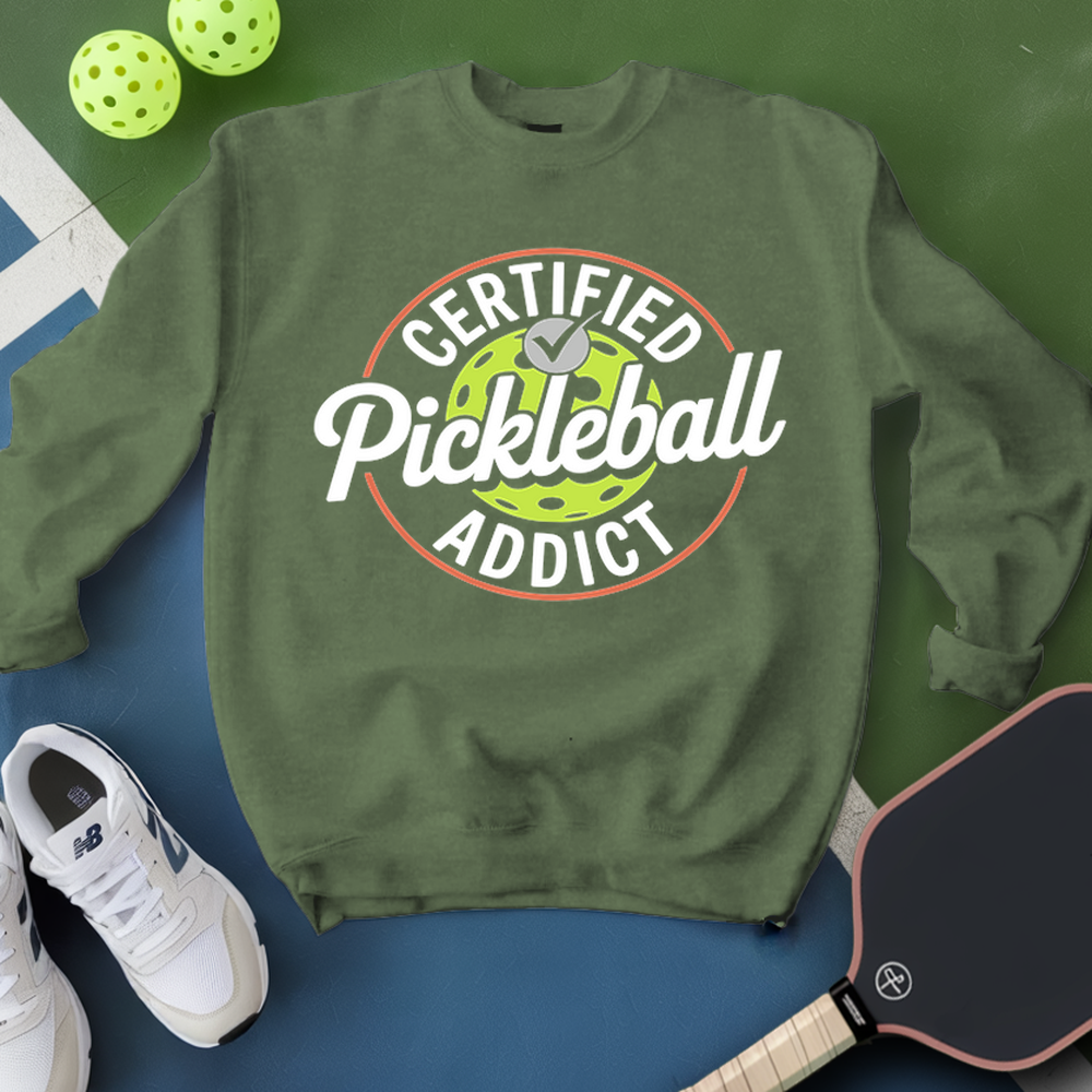 Green sweatshirt with 'Certified Pickleball Addict' text, pickleball paddles, and balls on a green and blue background