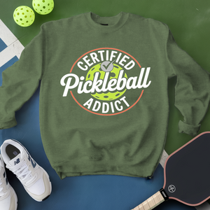 Green sweatshirt with 'Certified Pickleball Addict' text, pickleball paddles, and balls on a green and blue background