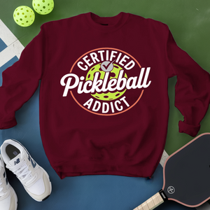 Maroon sweatshirt with 'Certified Pickleball Addict' text, pickleball paddle, and balls on a green and blue background