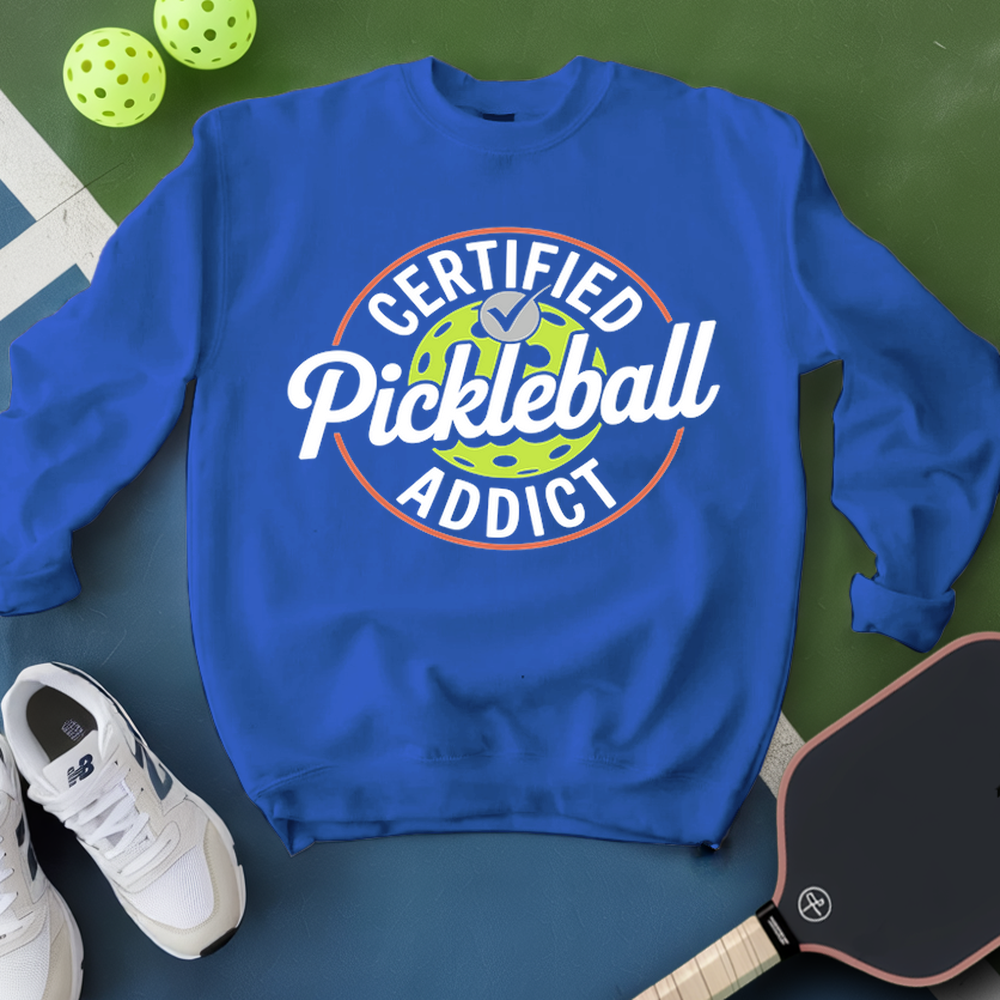 Blue sweatshirt with 'Certified Pickleball Addict' text on a green surface with pickleballs and a paddle.