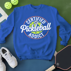 Blue sweatshirt with 'Certified Pickleball Addict' text on a green surface with pickleballs and a paddle.