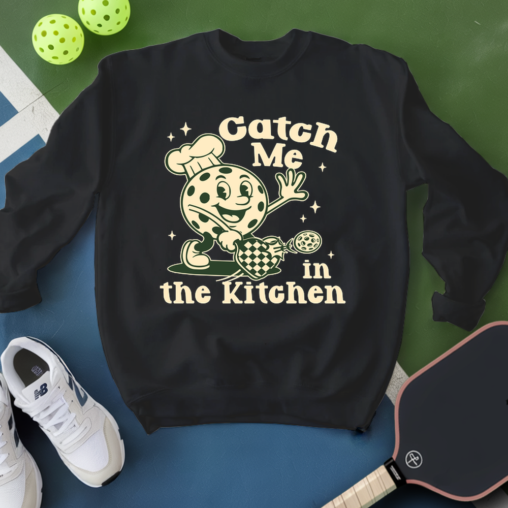 Black sweatshirt with a graphic design and text 'Catch Me in the Kitchen' on a blue and green background.
