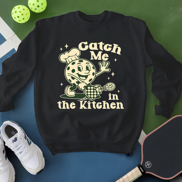 Black sweatshirt with a graphic design and text 'Catch Me in the Kitchen' on a blue and green background.