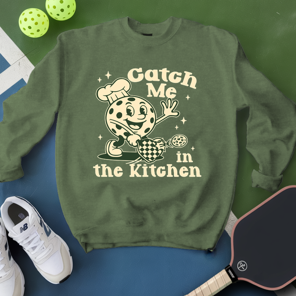 Green sweatshirt with cartoon character and text 'Catch Me in the Kitchen' on a blue and green background.