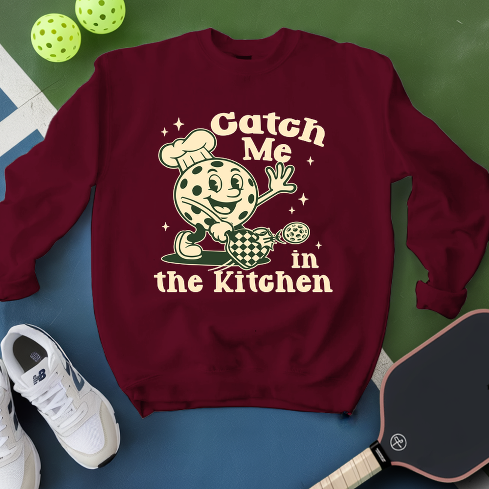 Maroon sweatshirt with cartoon character and text 'Catch Me in the Kitchen' on a green and blue background.