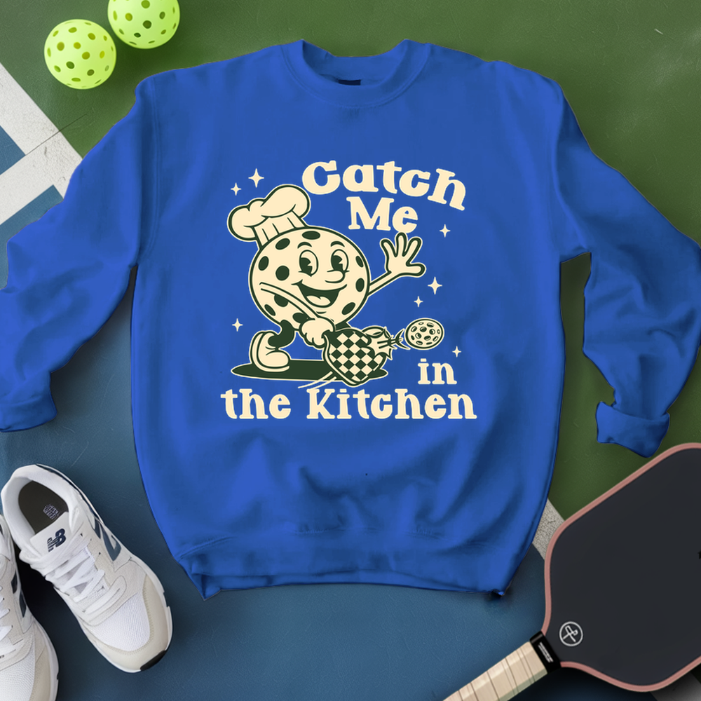 Blue sweatshirt with a cartoon character and text 'Catch Me in the Kitchen' on a green surface with pickleball equipment.