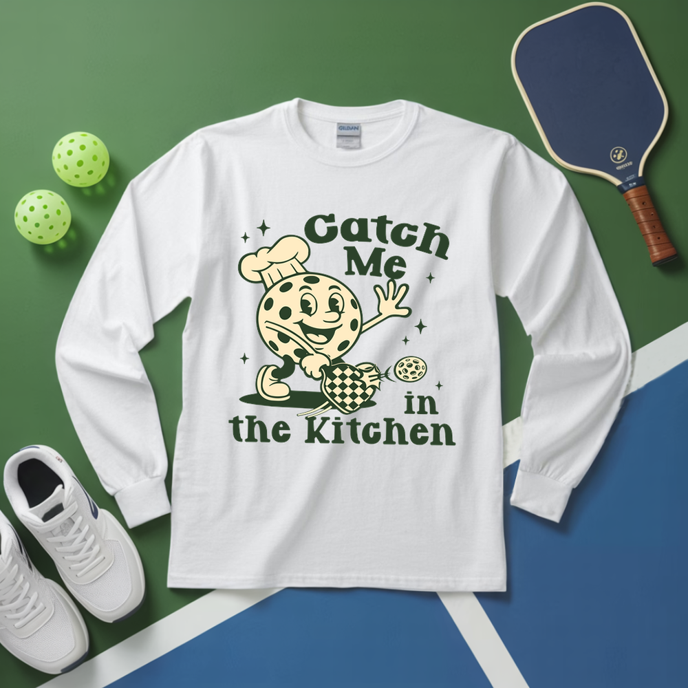 White long-sleeve shirt with a cookie graphic and text 'Catch Me in the Kitchen' on a green and blue background.